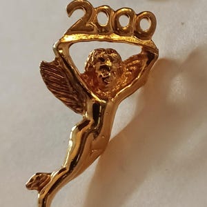 Puede incluir: Un broche dorado que representa un querubín sosteniendo el número "2000" sobre su cabeza. El querubín tiene alas y está en una pose elegante. El broche probablemente está hecho de metal y tiene un acabado brillante.