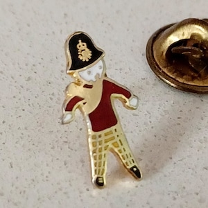 May include: A gold-colored pin of a cartoon soldier wearing a red and gold uniform, a black hat with a gold crown, and plaid pants. The pin has a gold back with a safety pin.