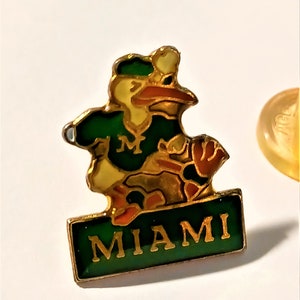 May include: A gold-colored pin with a cartoon mascot wearing a green and gold baseball uniform. The mascot is a bird with a green hat and a red beak. The pin has a green banner with the word "MIAMI" in gold letters.