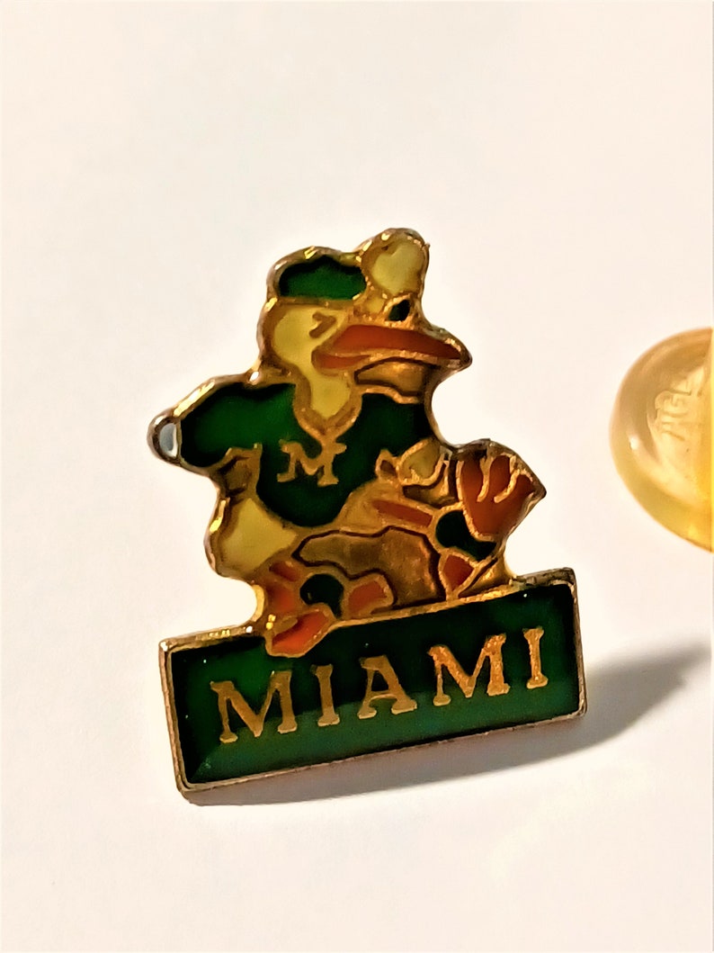 May include: A gold-colored pin with a cartoon mascot wearing a green and gold baseball uniform. The mascot is a bird with a green hat and a red beak. The pin has a green banner with the word "MIAMI" in gold letters.