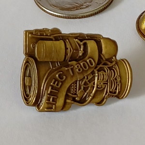 Puede incluir: Un pin de metal dorado que presenta una representación detallada de un motor con el texto "LHTEC T800" inscrito en el lateral.