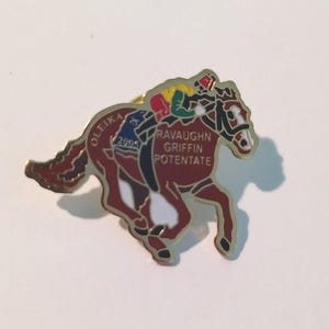 以下が含まれることがあります： 騎手を乗せて走る馬の形をした金属製のラペルピン。馬はゴールドの縁取りが施されたダークレッド。馬には「OLEIKA」「2005」「RAVAUGHN GRIFFIN POTENTATE」という文字が刻まれている。騎手は黄色と緑のシャツに赤い帽子を着用している。