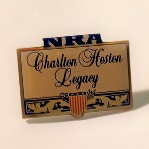 Puede incluir: Pin de metal dorado con un logotipo NRA Charlton Heston Legacy azul y blanco. El logotipo presenta un águila estilizada con un escudo en el centro.