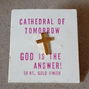 May include: A gold-colored cross pin on a white card. The card has the text "CATHEDRAL OF TOMORROW" and "GOD IS THE ANSWER!" in pink. The bottom of the card reads "18 KT. GOLD FINISH".