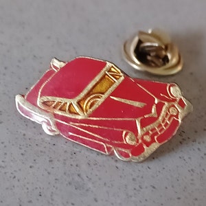 May include: A red vintage car pin with gold accents. The car has a chrome bumper and a detailed grill. The pin has a safety clasp on the back.