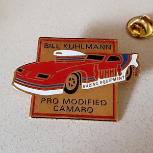 May include: A gold-colored pin featuring a red, white, and blue race car with the words "Bill Kuhlman" and "Pro Modified Camaro" on the front. The car has a white roof and the words "Summit Racing Equipment" on the side.