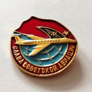 Pode incluir: Um broche metálico circular com um avião dourado a voar sobre uma secção de globo azul-claro. O fundo inclui um campo vermelho e um emblema de asa preta com uma estrela dourada. O texto cirílico "СЛАВА СОВЕТСКОЙ АВИАЦИИ" está gravado a ouro ao longo da margem inferior.