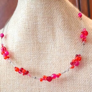 May include: A delicate silver necklace with pink and red beads arranged in clusters. The necklace is displayed on a fabric-covered mannequin.