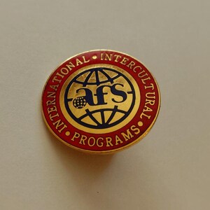 May include: A round pin with a gold border and a red outer ring. The pin features the text "INTERNATIONAL INTERCULTURAL PROGRAMS" in red. The center has a blue and gold globe design with the letters "afs" in blue.