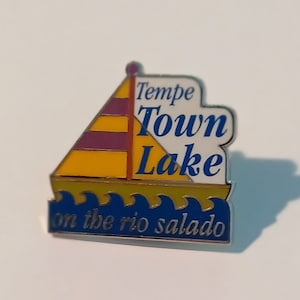 May include: A blue and yellow enamel pin with a sailboat design. The pin says "Tempe Town Lake on the rio salado".