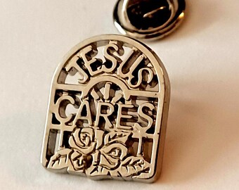 May include: Silver-toned lapel pin with the words "JESUS CARES" in an arched design. The pin features a floral pattern at the bottom and a separate pin back. The pin is likely made of metal.