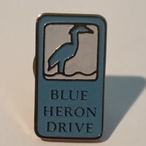 Op de afbeelding: Een rechthoekige speld met een blauw en wit ontwerp. De speld heeft een blauw reiger silhouet in een witte rechthoek, met de woorden "BLUE HERON DRIVE" in blauw eronder. De speld heeft een goudkleurige rand.