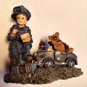 May include: A decorative figurine featuring a young boy in a navy blue uniform holding a paper, standing next to a vintage car with a teddy bear passenger. The base has the word "STOP" on blocks.