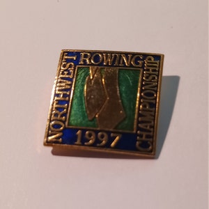 Puede incluir: Un pin de color dorado con un borde azul y remos de remo verdes y dorados. El texto "Northwest Rowing Championship 1997" está impreso en el pin.