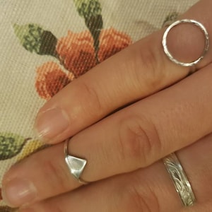 Puede incluir: Tres anillos de plata en un dedo. El anillo en el dedo índice es un círculo simple. El anillo en el dedo medio es de forma triangular. El anillo en el dedo anular es una banda delgada con un diseño texturizado.
