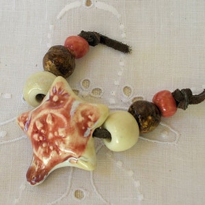 Peut inclure: Un pendentif en céramique en forme d'étoile avec un glaçage brun et rouge, enfilé sur un cordon en cuir brun avec des perles en céramique beige et brun.