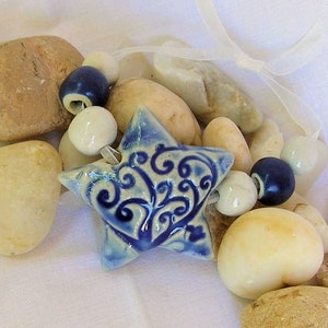 May include: A blue and white ceramic star with a swirling floral design hangs from a white ribbon with white and blue beads. The star is surrounded by smooth, light brown stones.