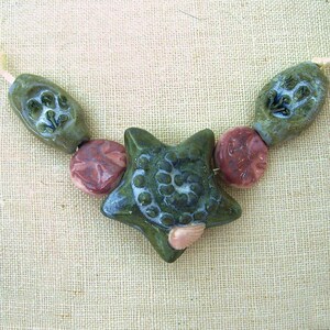Peut inclure: Un collier composé de perles en céramique vertes et roses. Les perles sont en forme d'étoiles, de cercles et de feuilles. Le collier est enfilé sur un cordon blanc.