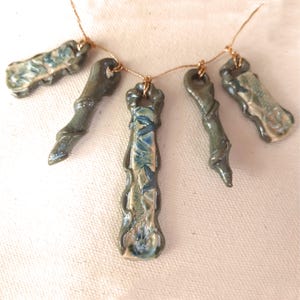 May include: A necklace with five ceramic pendants, each with a unique, organic shape and a blue and green glaze. The pendants are strung on a thin gold cord.