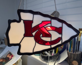 Op de afbeelding: Een pijlpunt van gekleurd glas met een rood "KC" logo. De pijlpunt hangt aan een zilveren ketting.