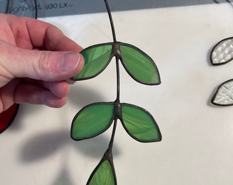 May include: A stained glass leaf design with a green and brown color scheme. The leaves are connected by a thin black wire.