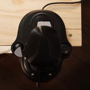 May include: A black joystick with a gray lever and a power switch is mounted on a light-colored wooden surface. The joystick has a rounded base and a central control stick. A black cable extends from the top.
