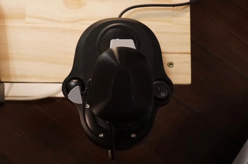 May include: A black joystick with a gray lever and a power switch is mounted on a light-colored wooden surface. The joystick has a rounded base and a central control stick. A black cable extends from the top.