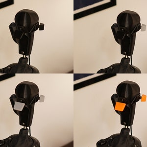 May include: A black, articulated robot head with a rounded top and various attachments. The attachments are shown in black, gray, and orange. The robot head is mounted on a black stand, with a white wall in the background.