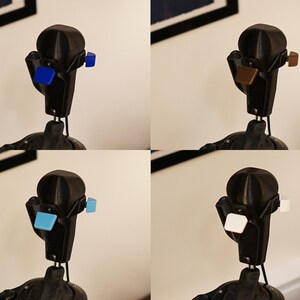 May include: A black 3D-printed robot head with four different colored square attachments: blue, brown, light blue, and white. The robot head has a rounded top and a rectangular body with a thin neck.
