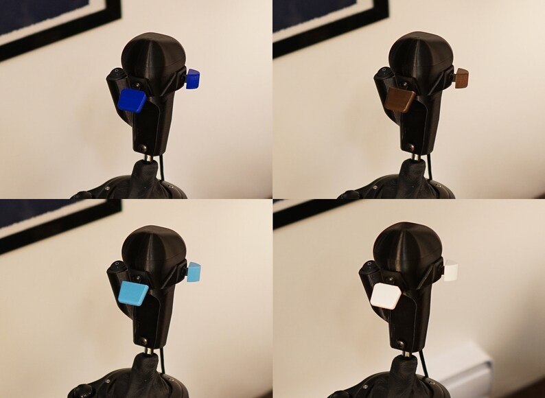 May include: A black 3D-printed robot head with four different colored square attachments: blue, brown, light blue, and white. The robot head has a rounded top and a rectangular body with a thin neck.