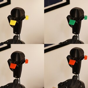 May include: A black joystick with interchangeable colored buttons: yellow, green, and orange. The joystick is mounted on a black base and has a unique, angular design. The image is a composite of four different views of the same joystick.