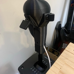 May include: Black gaming gear shift with a rounded top and multiple buttons. The gear shift is mounted on a black base with numbered gear positions. A white cable is connected to the gear shift.