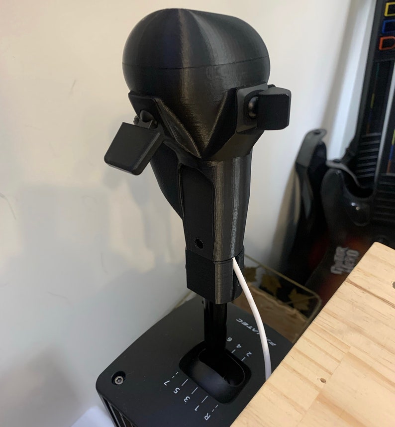 May include: Black gaming gear shift with a rounded top and multiple buttons. The gear shift is mounted on a black base with numbered gear positions. A white cable is connected to the gear shift.