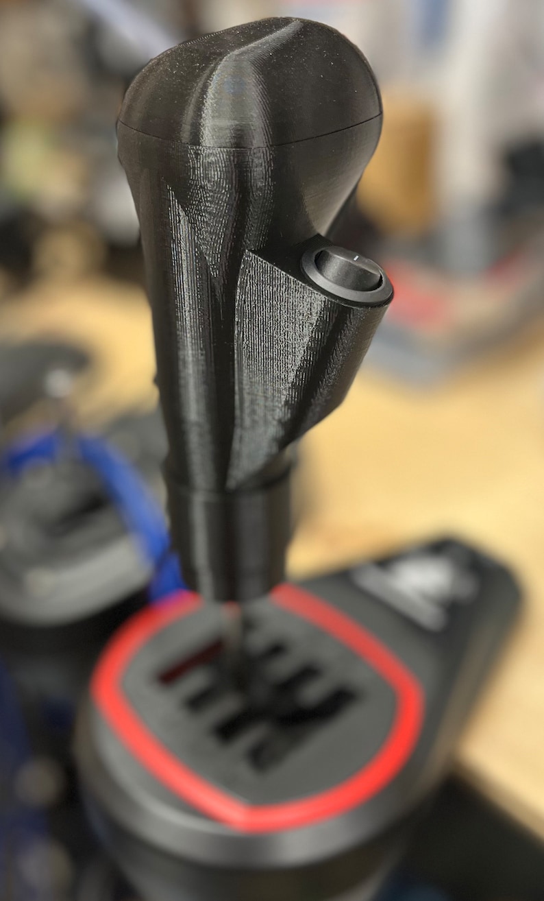 May include: Black gear shift knob with a textured surface and a small button. The knob is attached to a black and gray gear shift pattern with a red border. Automotive accessory.