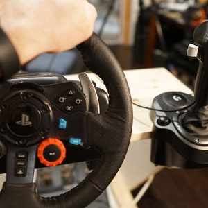 May include: Black and gray gaming steering wheel with multiple buttons and an orange dial. A black joystick is connected to the wheel. The wheel is being held by a person wearing a black watch.