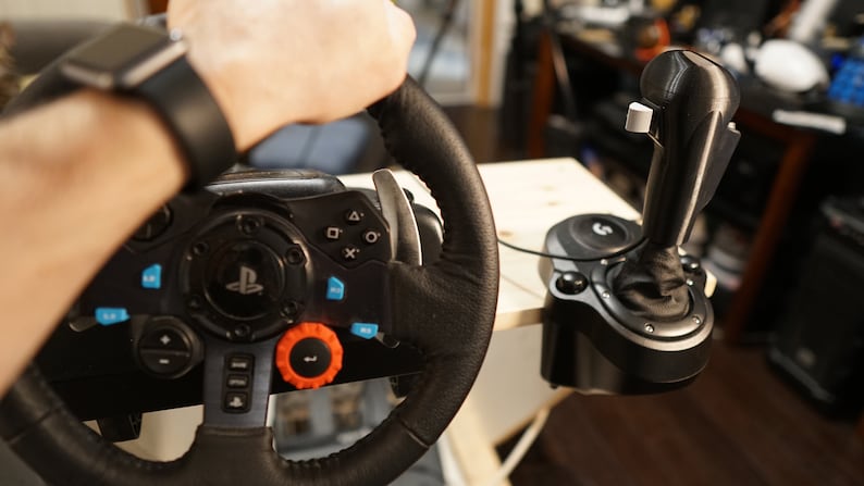 May include: Black and gray gaming steering wheel with multiple buttons and an orange dial. A black joystick is connected to the wheel. The wheel is being held by a person wearing a black watch.
