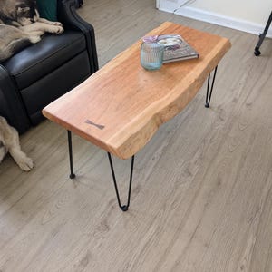 May include: A live edge coffee table with a natural wood grain finish. The table has black hairpin legs and a small wooden butterfly joint. A glass and a book are on the table.