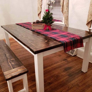 May include: A dark brown wooden farmhouse table with white legs and a red and black plaid table runner. A small wooden bench is in the foreground.