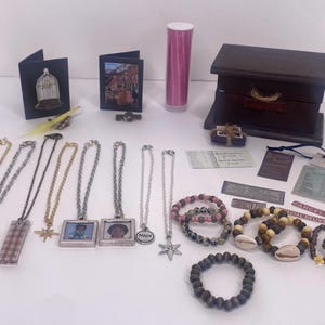 May include: A collection of jewellery and accessories, including necklaces with various pendants, bracelets, and a small wooden chest. Other items include greeting cards, a pink cylinder, and decorative pins. The jewellery features heart, star, and photo pendants.
