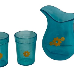May include: A turquoise jug and two matching glasses. The jug has a handle and a flower design. The glasses and jug have a flower design. The set is made of a transparent material.