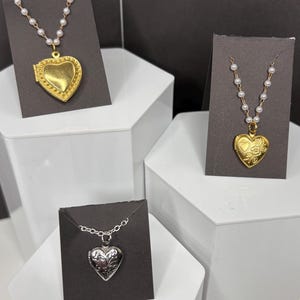 May include: Three heart-shaped locket necklaces on display. One is gold-toned with a pearl chain, another is silver-toned, and the third is gold-toned with a pearl chain. The lockets are displayed on black cards.