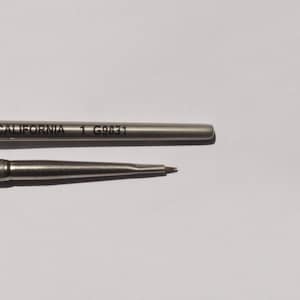 May include: Close-up of two silver-colored pen refills. The top refill has the text "CALIFORNIA 1 G9831" printed on it. The bottom refill has a fine, brown tip. Both refills are on a white background.