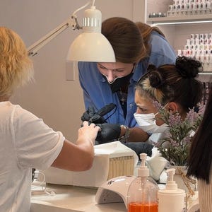 May include: A nail salon scene with three people. One person's hand is being worked on by a person wearing black gloves. A lamp is positioned over the hand. Bottles of liquid and a UV nail lamp are on the table. Shelves of nail polish are in the background.