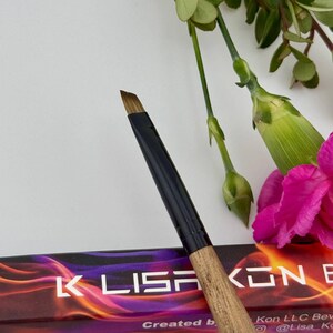 May include: A makeup brush with a black handle and a wooden grip. The brush head is angled and has brown bristles. The background includes pink flowers and green foliage. The text "K Lisa Kon B" is visible.