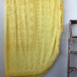 May include: A vibrant yellow bedspread or throw blanket with a vintage design featuring floral and geometric patterns. The edges are finished with a fringe. The textile is hanging on a wooden rod against a neutral wall.