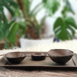 Può includere: Tre ciotole in ceramica marrone di diverse dimensioni su un vassoio rettangolare. Le ciotole hanno un aspetto rustico e strutturato con bordi irregolari. Il vassoio è di un colore oro tenue. Lo sfondo è sfocato.