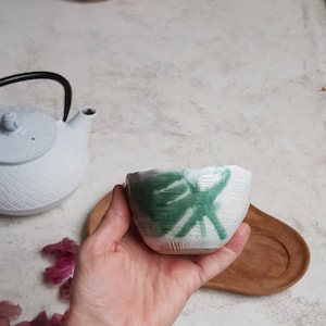 May include: A white ceramic tea bowl with a textured surface and a green abstract design. A white teapot with a black handle and a wooden tray are in the background. The bowl is held in a hand.