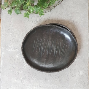 May include: Two handmade, dark grey ceramic plates. Each plate has a slightly irregular, round shape with a textured rim. The surface of each plate features a series of vertical, etched lines, adding a unique design element.