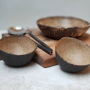 May include: A set of handmade ceramic bowls with a speckled interior and dark exterior. The bowls are various sizes and shapes, with a larger bowl in the background and two smaller bowls in the foreground. Chopsticks and a spoon rest on a wooden surface.