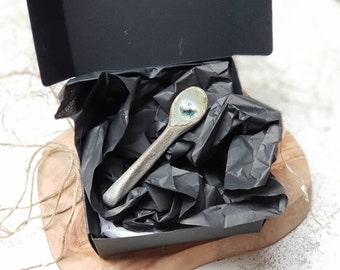 May include: A handmade ceramic spoon with a speckled, metallic finish and a light blue and green bowl. The spoon rests in a black gift box with black tissue paper, presented on a wooden board.